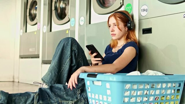 Young redhead girl sitting on the floor of a laundry room next to a basket of clothes listening to music with her smart phone waiting for the clothes to be washed