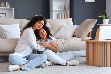 Fototapeta premium Young African-American woman with her daughter sitting on floor and hugging at home