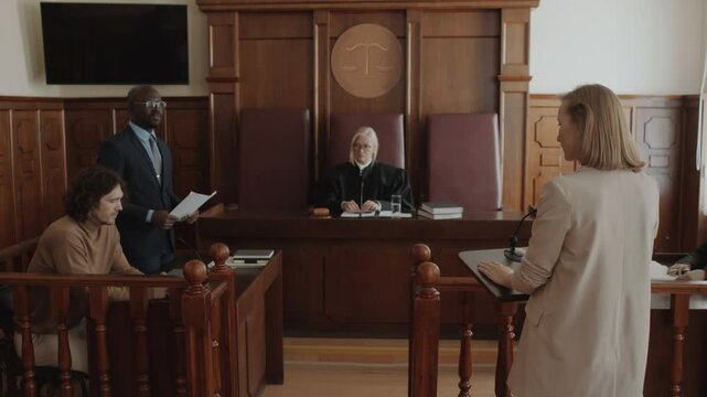 Black male attorney asking Caucasian female witness questions about suspect sitting near him during trial in courtroom, judge listening