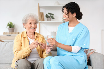 Young African-American female medical worker giving elderly woman pills and glass of water in nursing home