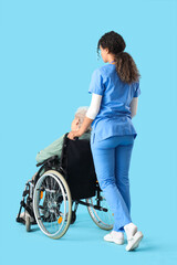 Young African-American female medical worker with elderly woman on wheelchair against blue background