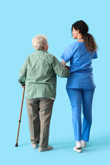 Young African-American female medical worker hugging elderly woman on blue background, back view