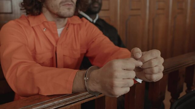 Close up of hands of young male suspect in metallic cuffs wearing orange jumpsuit sitting near policeman at court trial