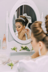 A woman is getting ready for a wedding and is putting on her jewelry. The mirror reflects her as...