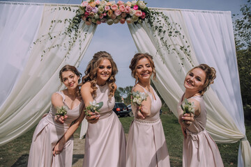Four women are posing for a picture in front of a white archway. They are holding flowers in their...