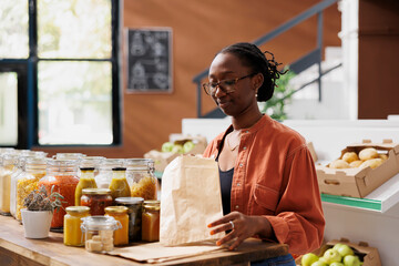 Black woman at wooden counter holding paper bag for putting fresh bio food product at convenience store. African american customer grasping small brown sack for freshly harvested items at local shop.