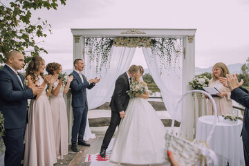 A bride and groom are kissing in front of a white arch. The bride is wearing a white dress and the...