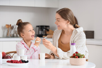 Cute little girl in pajamas with her mom eating yogurt in kitchen