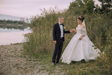 A bride and groom are walking on a beach near a body of water. The bride is wearing a white dress and the groom is wearing a suit. Scene is romantic and happy