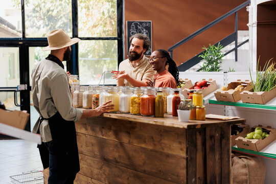 Male vendor offering advice to clients who are shopping for sustainable merchandise at eco friendly store. Owner of local supermarket describing food items in jars to the young multiracial couple.