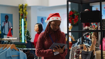 Obraz premium Employee wearing festive Santa hat crosschecking online shop clothing prices with physical store ones, looking to correct mismatches. Worker in Christmas decorated fashion boutique