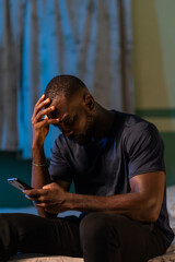 A young African man sits on a bed, holding his phone and looking down with a thoughtful expression....