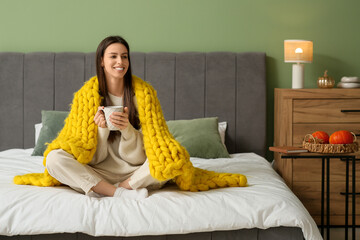 Beautiful young woman with warm yellow plaid and cup of tea sitting on bed in bedroom