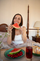 Lovely smiling woman enjoys a slice of watermelon in bed. A breakfast tray with raspberries, juice,...