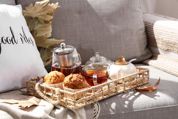 Wicker tray with teapots, cup of hot beverage, cupcakes and autumn leaves on sofa in living room