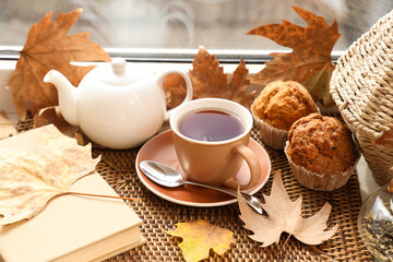 Teapot with cup of hot beverage, tasty cupcakes, book and autumn leaves on windowsill in room