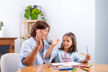 Fototapeta premium Grandmother giving high five to girl as she holds colored pencil while drawing at home. The scene captures moment of encouragement and admiration as the child engages in her creative activity 