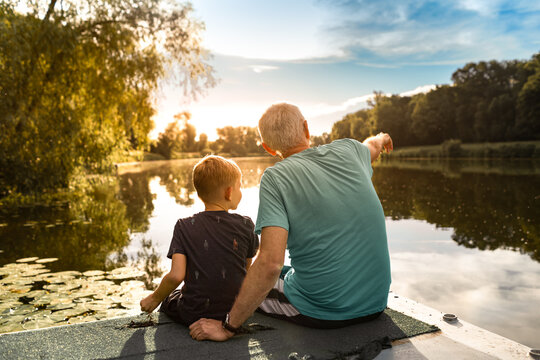 Grandfather and grandson sitting together outdoors on a dock by a lake bonding  enjoying nature sunset.  
