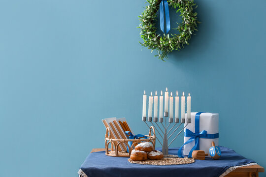 Menorah with donuts, books, dreidels and gift for Hannukah on table near blue wall