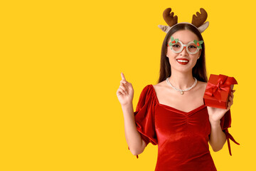 Beautiful young Asian woman in Christmas deer horns and glasses with gift box on yellow background