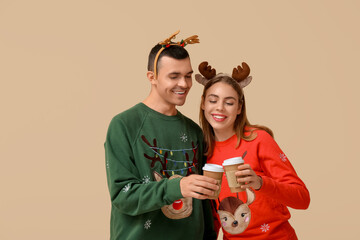 Beautiful young couple in reindeer horns with paper cups of coffee on beige background