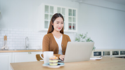 Beautiful young Asian woman working with laptop computer in home kitchen. Work at home