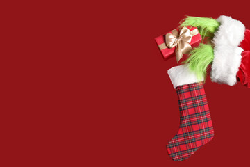 Green hairy hands of creature in Santa costume with sock and Christmas gift box on red background