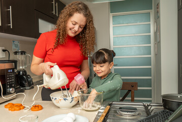 Mom blending dough with a hand mixer while her happy daughter adds flour with her hands, smiling as they bake cookies together at home.