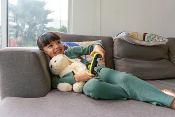 Obraz premium little girl playing video games lying on the sofa, happy kid smiling enjoying entertainment with technology