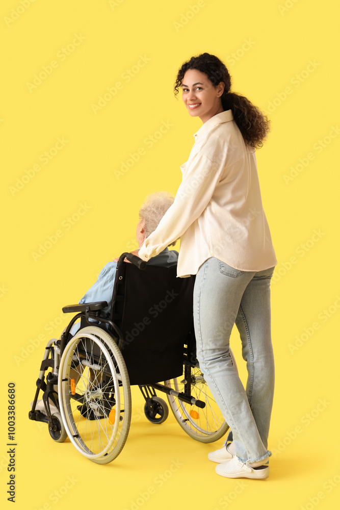 Wall mural Young African-American female medical worker with elderly woman on wheelchair against yellow background