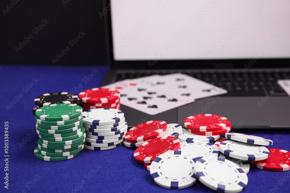 Poster Laptop, poker chips and playing cards on blue table, closeup. Online game