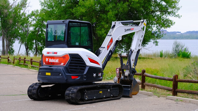 Bobcat mini excavator parked on paved area near a wooden fence overlooking a lake with trees in the background. Construction, public works, heavy equipment rental, landscaping services.