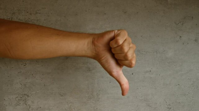 Man showing a thumbs down gesture in front of a concrete wall, conveying disapproval and negativity in a neutral setting