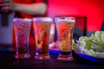 A cold beer glass sits on a cluttered desk during a late-night party, its golden hue reflecting the...