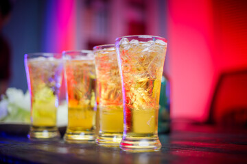 A cold beer glass sits on a cluttered desk during a late-night party, its golden hue reflecting the...