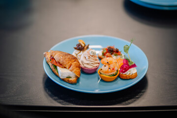 A plate of assorted gourmet appetizers, including a croissant sandwich, shrimp, canapés, and cream cheese bites on a blue plate..