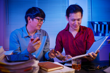 Two Asian businessmen work late into the night, sitting at a desk cluttered with a stack of papers...