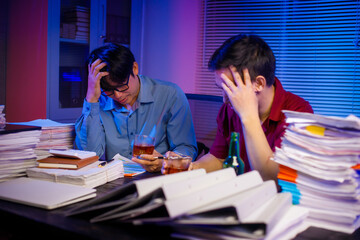 Two Asian businessmen work late into the night, sitting at a desk cluttered with a stack of papers...