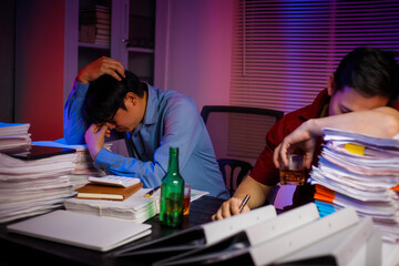 Two Asian businessmen work late into the night, sitting at a desk cluttered with a stack of papers...