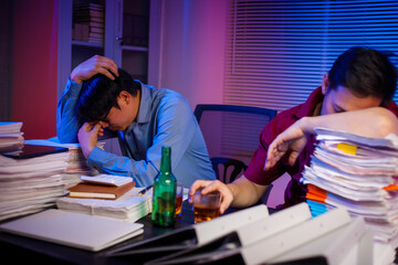 Two Asian businessmen work late into the night, sitting at a desk cluttered with a stack of papers...