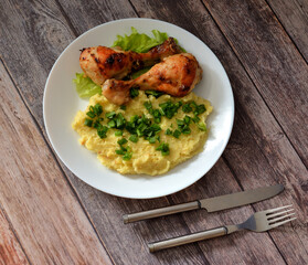Two baked chicken legs, mashed potatoes with chopped green onions on a wooden table, cutlery nearby.