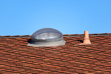 A residential roof covered by red asphalt shingles with a dome shaped skylight and a small vent pipe. Close up