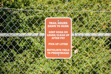 A trail information sign hangs on a chain-link fence, outlining guidelines for trail hours, pet...
