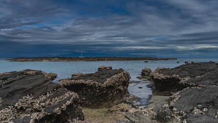 Littoral at low tide. Many attached clam shells can be seen on the exposed picturesque boulders. The yacht is far away, in the turquoise ocean. A tall mast against the sky and clouds. Madagascar.