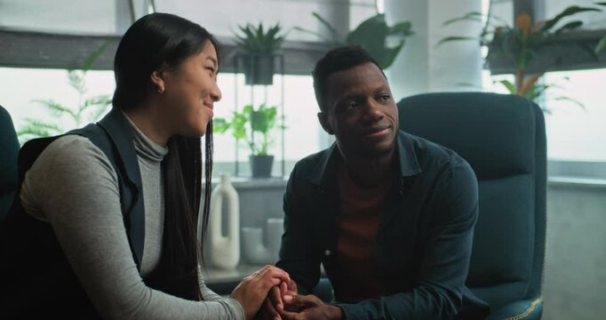 Tender couple sit on chairs holding hands, smile, find compromise after quarrel on couples therapy with therapist. Psychologist talks to multiethnic young family on counseling session. Psychology.