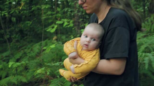 A woman wearing glasses and a black T-shirt is walking through a forest with a baby in her arms. View from the side. The camera is moving parallel to the woman. Slow motion. Evening walk