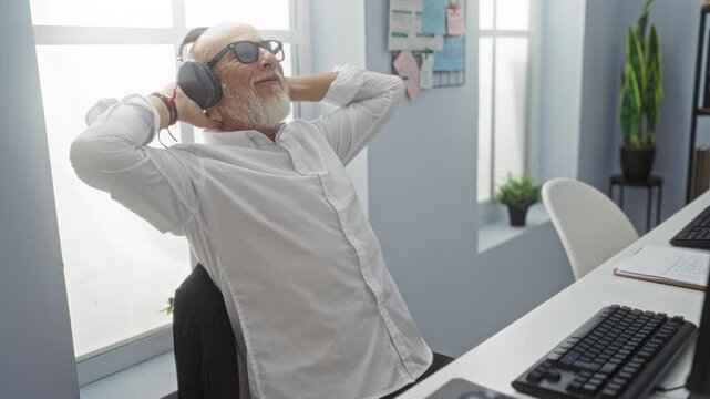 Middle-aged man with headphones relaxing at workplace indoors, wearing white shirt and glasses, sitting at desk in modern office with plants and windows in background.