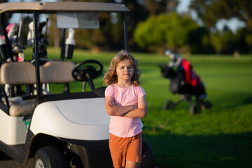 Fototapeta premium Kid in golf park near golf cart. Child summer vacation. Portrait of kid in spring park outdoors. Close-up face child playing outdoors in summer park.