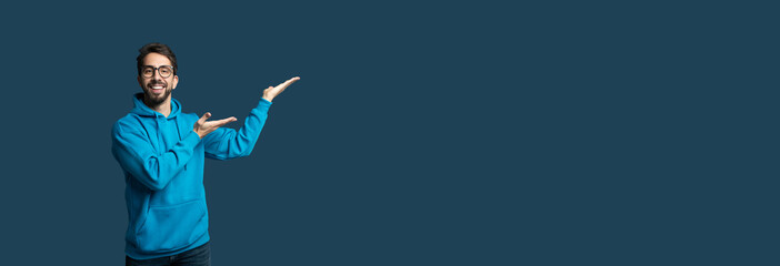 A man wearing a vibrant blue hoodie smiles and gestures with his hands, inviting viewer engagement against a solid blue background, panorama with copy space
