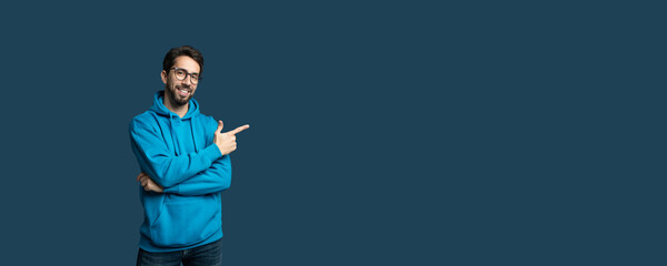 A young man wearing a blue hoodie stands against a solid dark background, smiling and pointing to...